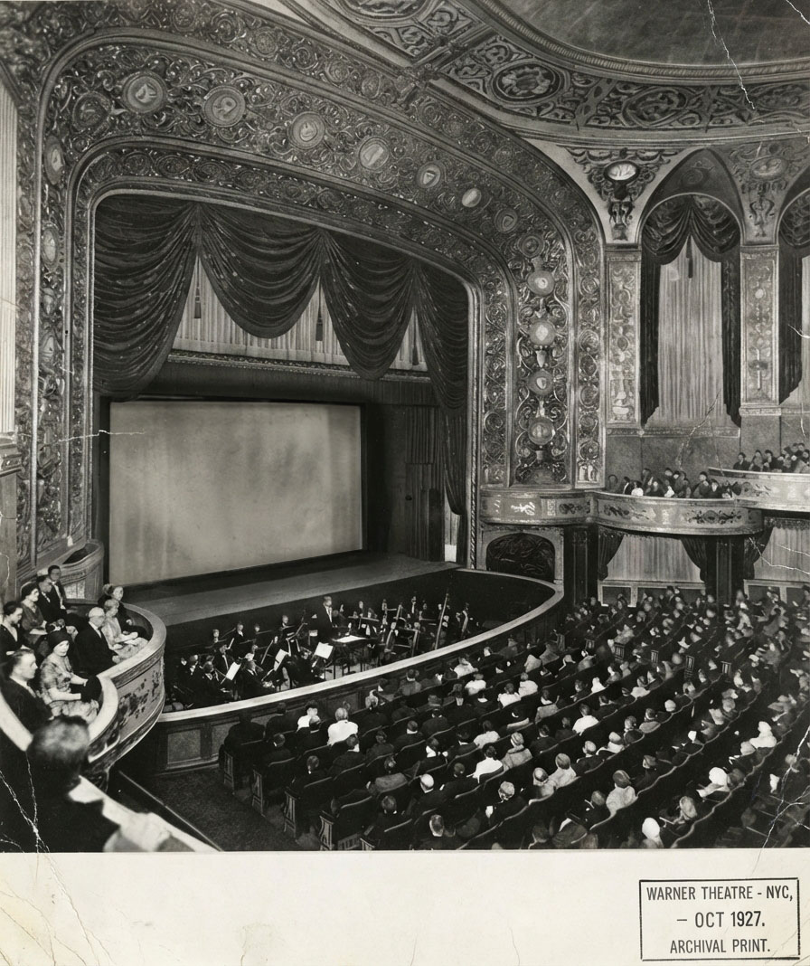 Warner Theatre, New York City, Oktober 1927. Im Orchestergraben sitzen die Musiker:innen vor der Leinwand.